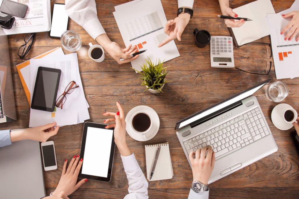 Draufsicht auf einen Tisch, wo gerade ein Meeting stattfindet und die Beteiligten Laptops, Handys und Dokumente benutzen.